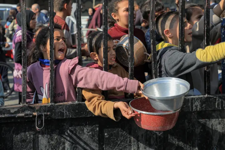 “Small boy reaching through a fence with a bowl in hand, asking for food from a crowded area”